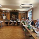 12 men and 1 woman with white nameplates in front of them, sitting around a "U-shaped" meeting table.